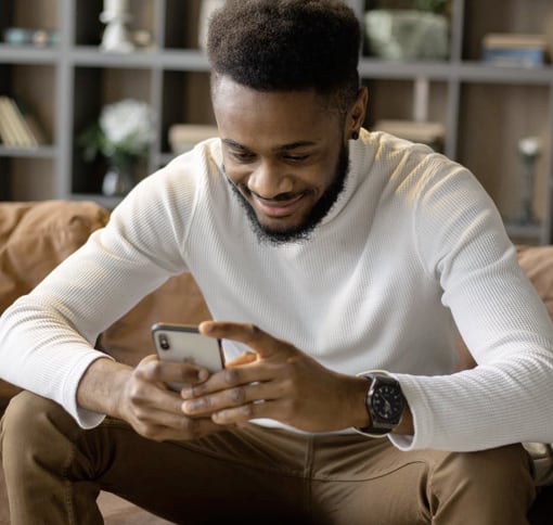 
              a man sitting on the couch in his living room smiling while making a video call with meet on his smartphone.
              The man is wearing dark brown pants and a white long-sleeved shirt, on his left wrist he has a watch.
              In the background out of focus you can see some modern style shelves with various objects on them.