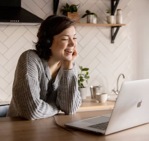 
              A smiling woman in her kitchen, on the kitchen island, making a video call with her laptop.
              The woman has brown hair that reaches the base of her neck and is wearing a gray wool sweater.
              In the background you can see the rest of the kitchen, with white majolica, the black hood,
              a shelf to the right of the hood with ornamental plants on it. You can also see the sink, which next to it,
              on the left, has two other small ones with ornamental plants.