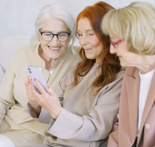 
              Of three adult women sitting next to each other while they are on a video call with a smartphone.
              The woman on the left is smiling, has graying white hair, wears eyeglasses and a beige shirt.
              The central woman who is the woman holding the smartphone in her hands instead,
              has long red hair that falls in front of her shoulders,
              you can see red nail polish on her fingernails and she is wearing a gray long-sleeved shirt.
              The last woman, the one sitting on the right, is framed in profile, has blonde bob hair,
              wears red eyeglasses and is wearing a white shirt under an elegant light brown jacket.